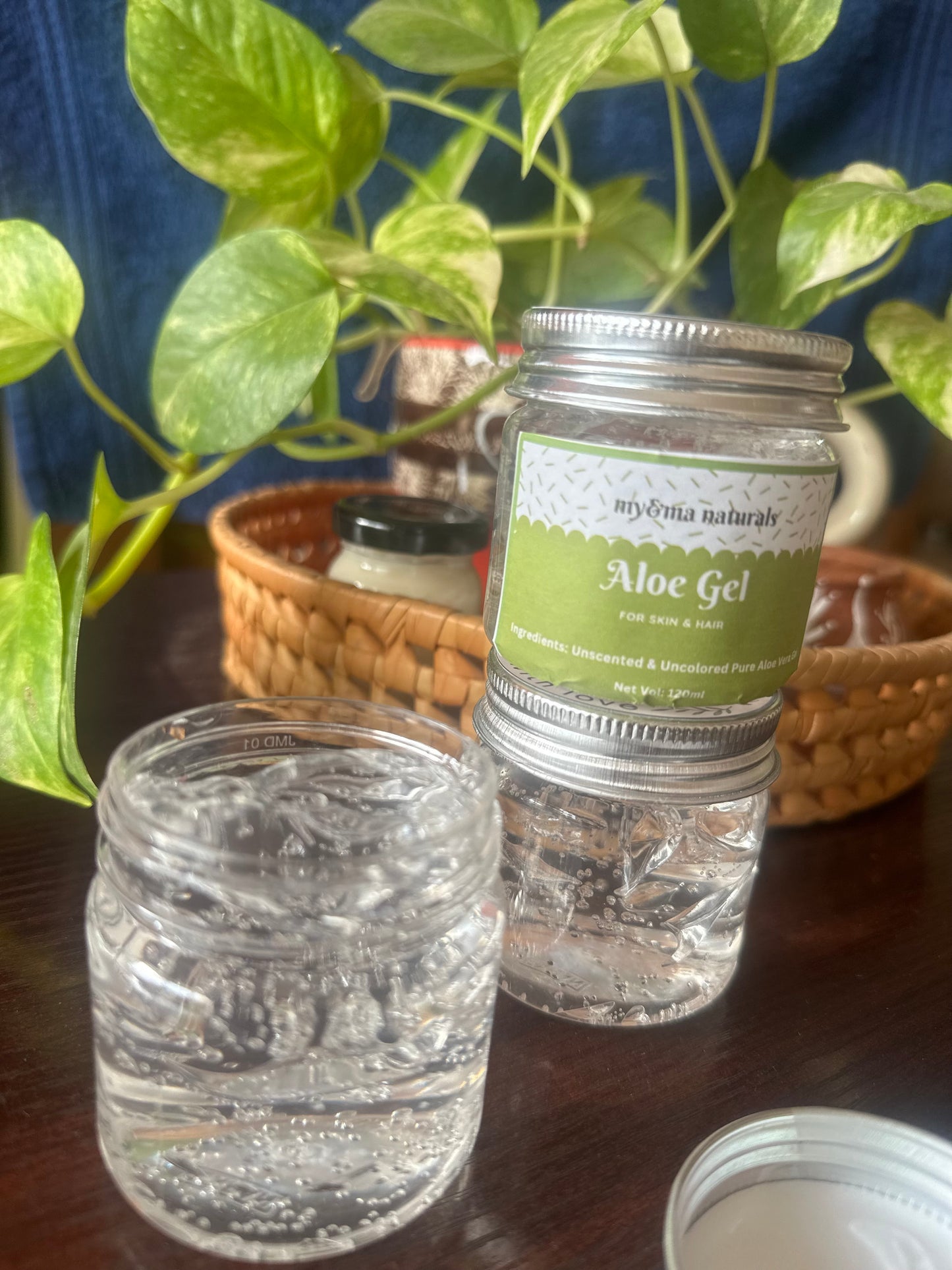 Aloe Vera Gel (100gm) in clear jars with a green label, surrounded by lush green plants and a wicker basket, showcasing its pure and natural composition for soothing skin, moisturizing, fighting skin aging, reducing blemishes, and promoting healthy hair.