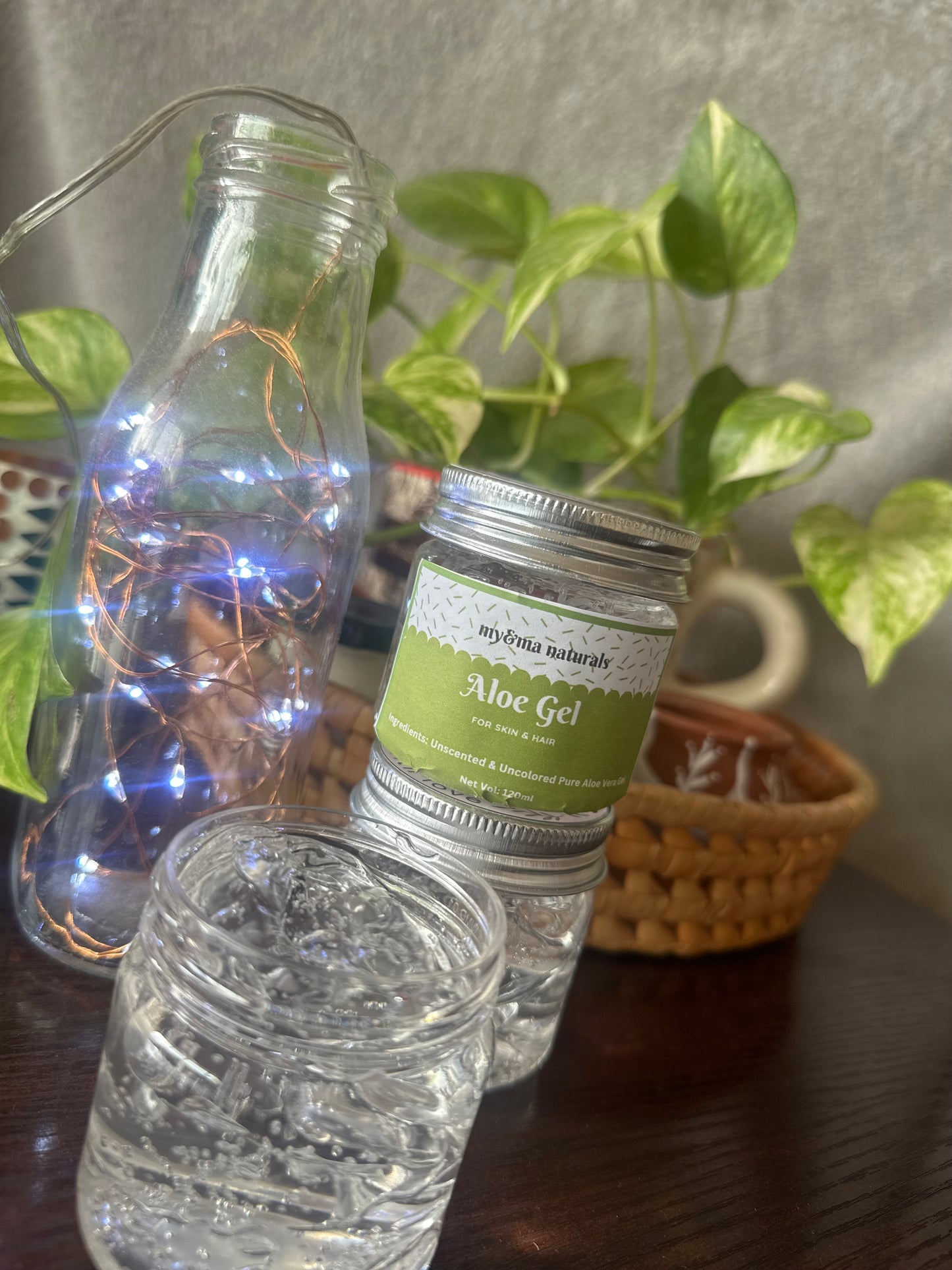 A beautifully presented 100gm jar of Aloe Vera Gel from 'My Byna Naturals', shown with an open and closed container against a cozy backdrop with plants and fairy lights.