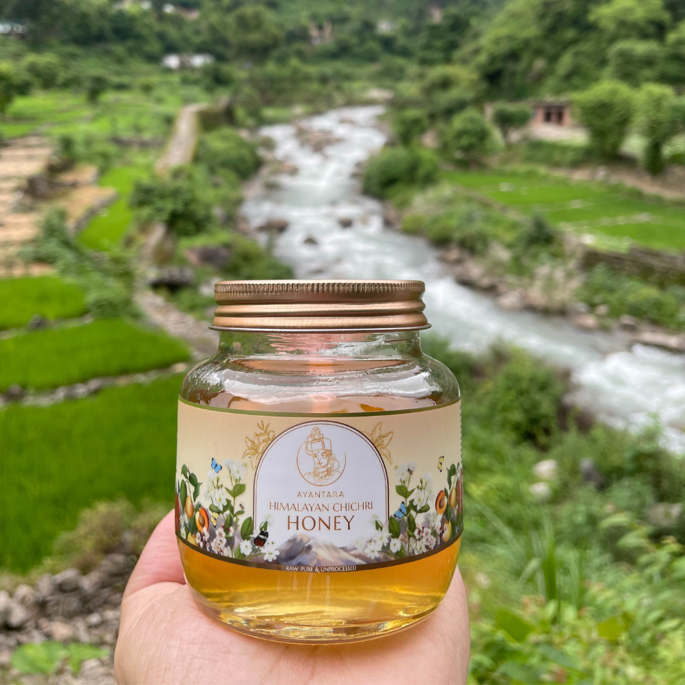 Close-up of Ayantara Himalayan Chichri Honey (250g) jar, held against a backdrop of lush green fields and a flowing river, showcasing the purity and raw essence of unprocessed, natural honey straight from the Himalayan region.
