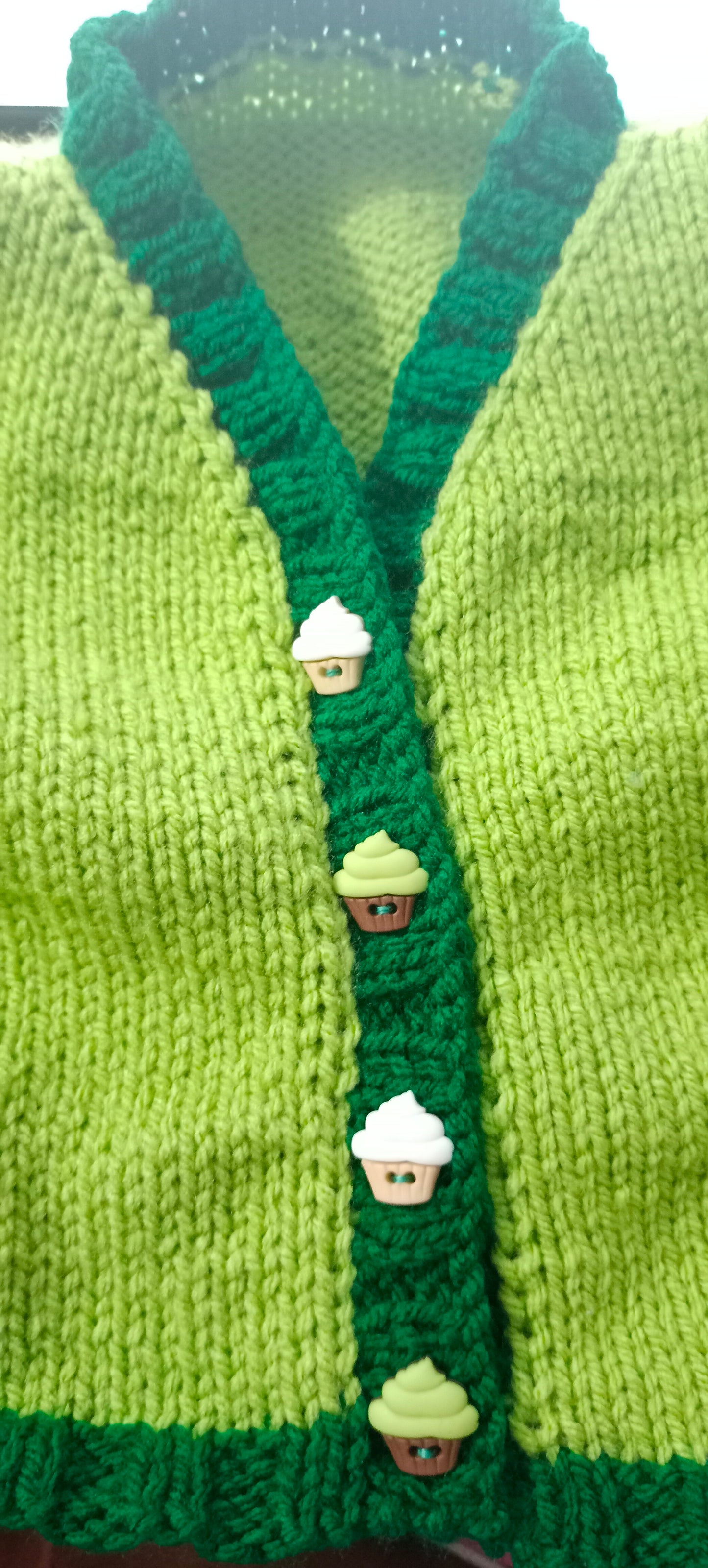 
                  
                    Handknitted light green v-neck toddler cardigan with dark green ribbing, adorned with adorable cupcake buttons, perfect for ages 6-9 months. Made from 100% acrylic yarn in a pet-free home.
                  
                