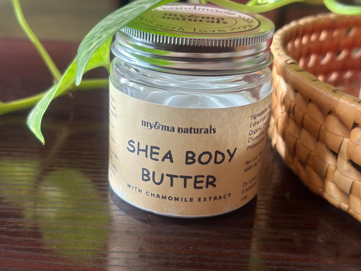 
                  
                    Close-up view of my&ma naturals Shea Body Butter (100gm) with chamomile extract in a recyclable glass jar, placed beside a leafy plant and a woven basket on a wooden table.
                  
                