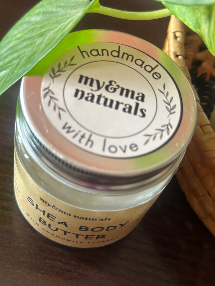 
                  
                    my&ma naturals Shea Body Butter (100gm) in a glass jar with a handmade label, placed next to a green plant on a wooden surface.
                  
                