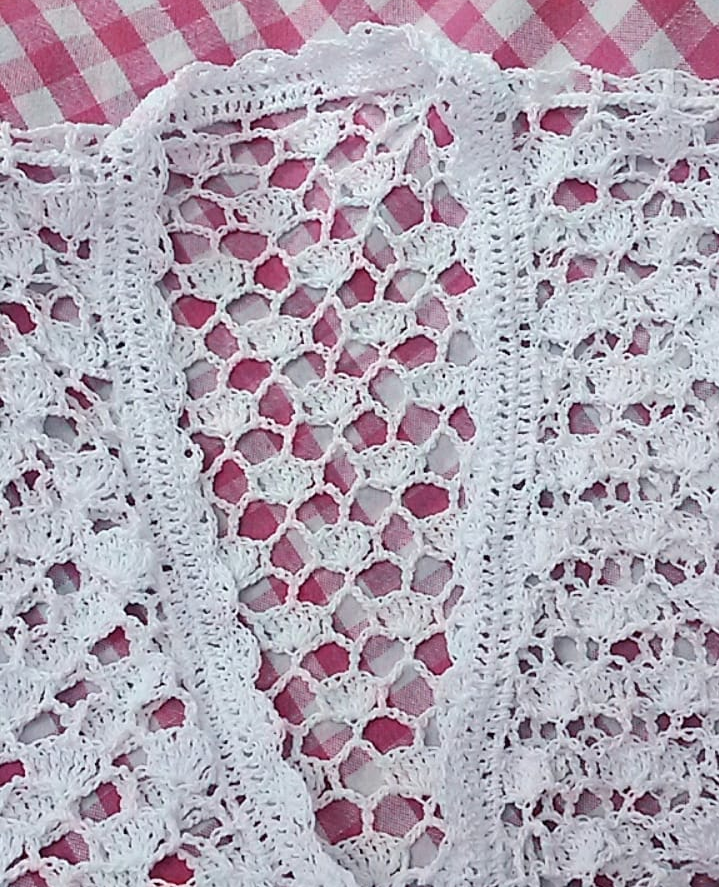 
                  
                    Close-up of a white lace beach cover-up made from 100% cotton with intricate crochet patterns, displayed on a pink and white checkered background. Perfect for adding a touch of elegance to your summer beachwear or as a stylish cardigan.
                  
                