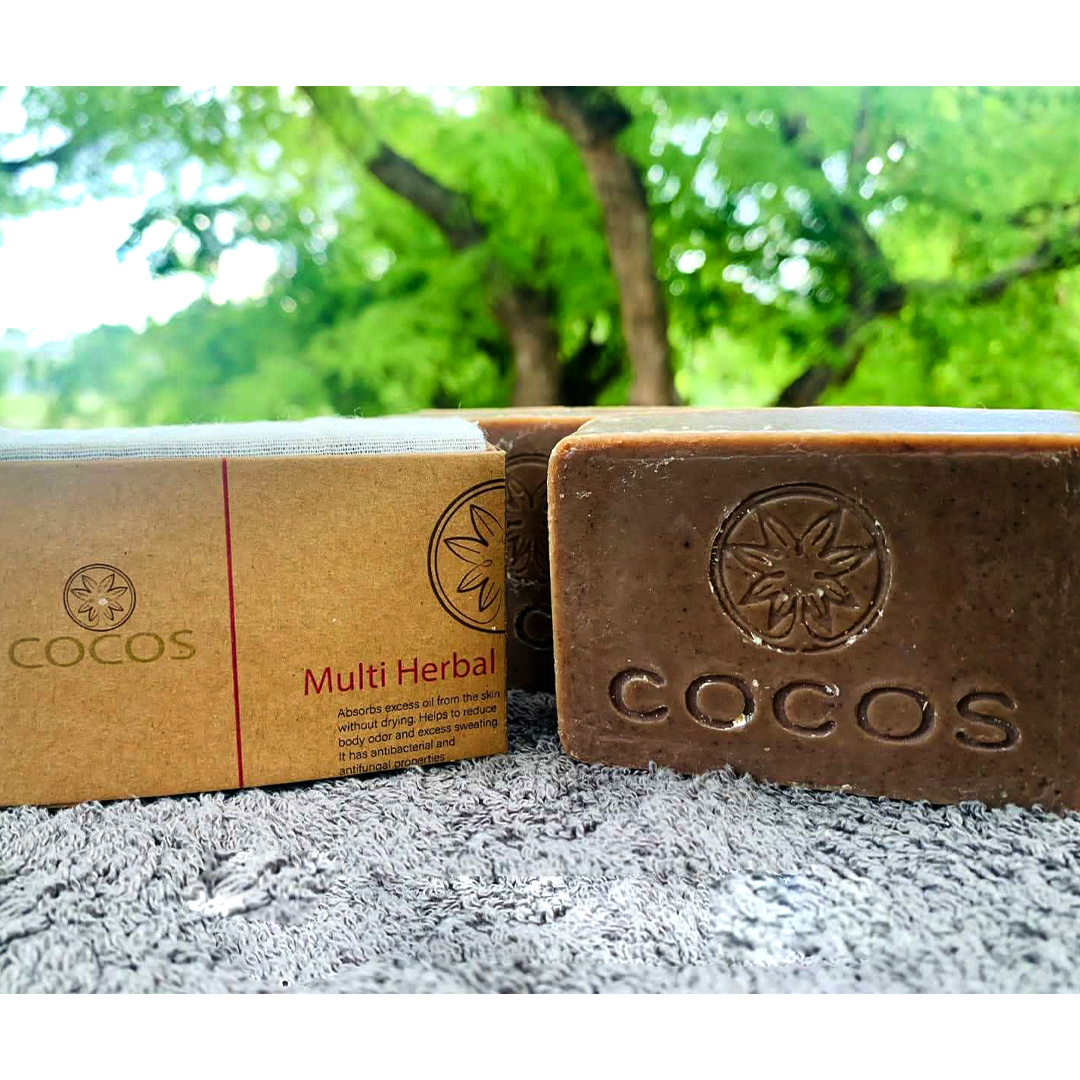 "Invigorating Multi Herbal Soap by COCOS, shown nestled on a soft towel with lush greenery in the background, ready to cleanse and refresh your skin while reducing body odor and excess sweating."